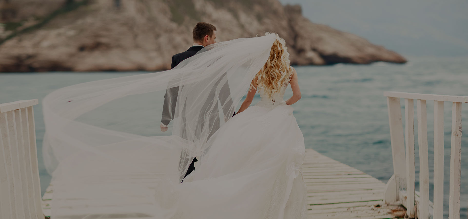 Bruid met bruidskapsel van blond los haar met krullen en sluier wapperend in de wind loopt hand in hand met bruidegom op steiger in de zee.