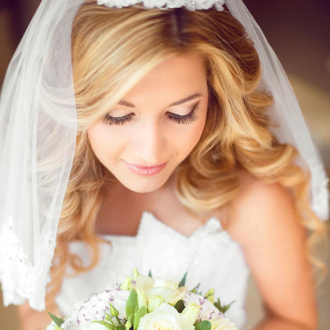 Bruid met sluier en bruidskapsel van los blond haar met slagen, krullen en lok kijkt naar haar bruidsboeket.